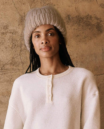 A person wearing The Handknit Chunky Beanie in cream by THE GREAT. HOL 24 PLUSH KNITS stands against a textured wall, dressed in a white sweater.