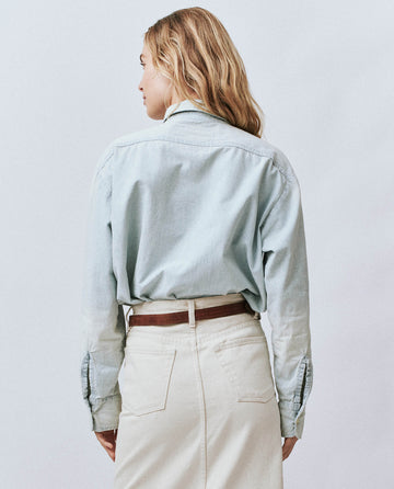 A woman with blonde hair is wearing an oversized boyish light blue denim shirt, known as The Road Shirt in Riverbed Wash from THE GREAT. FALL 24 COLLECTORS collection, paired with a beige skirt, standing with her back to the camera.