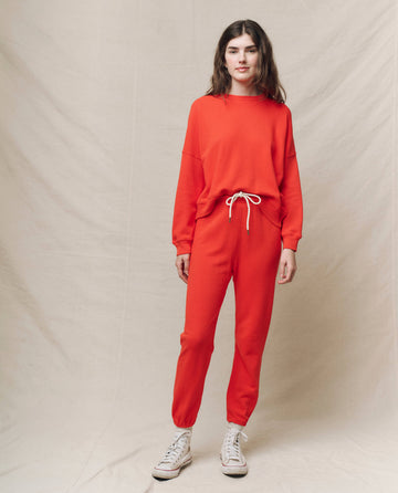 A person wears The Teammate Sweatshirt in Tart by THE GREAT. SP25 KNITS and matches it with joggers, posing against a neutral backdrop in team apparel style, complemented by crisp white sneakers.