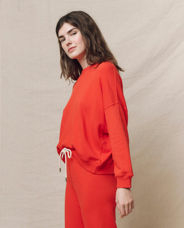 Clad in a bold red "The Teammate Sweatshirt. Solid -- Tart" by THE GREAT. SP25 KNITS, paired with matching pants, a person stands confidently against a neutral backdrop.