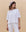 A woman wears THE GREAT. PF25 HEAVY JERSEY KNITS' The Track Crew. -- True White, paired with white pants and a patterned headscarf, standing against a beige background in an oversized, vintage athletic-inspired silhouette.