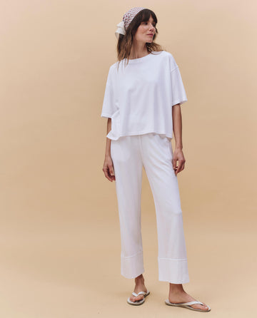 A woman stands against a beige background wearing THE GREAT. PF25 HEAVY JERSEY KNITS The Jersey Win Pant in True White, a loose heavyweight jersey white t-shirt, white flip-flops, and a patterned headscarf.