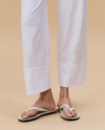 Wearing THE GREAT. PF25 HEAVY JERSEY KNITS The Jersey Win Pant in True White with beige flip-flops, only the lower legs and feet appear on a light beige surface—ideal loungewear for relaxed days.