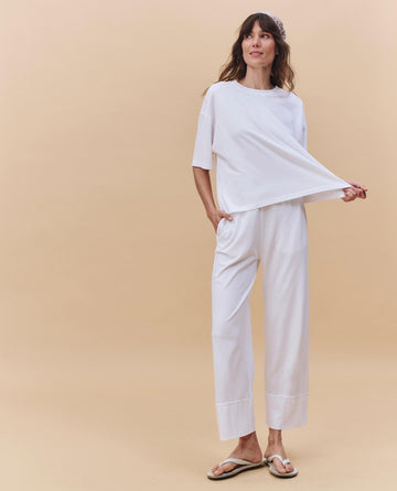 A woman stands against a beige background wearing THE GREAT. PF25 HEAVY JERSEY KNITS The Track Crew. in True White, styled with white wide-leg pants, flip flops, a headband, and holding out the side of her shirt.
