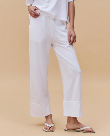 A person wears THE GREAT. PF25 HEAVY JERSEY KNITS’ The Jersey Win Pant in True White, paired with a matching white top and flip-flops, standing against a plain beige background.