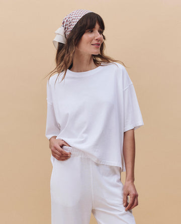 A woman wears THE GREAT. PF25 HEAVY JERSEY KNITS The Track Crew. -- True White, styled with white pants and a patterned headscarf, standing against a beige background.