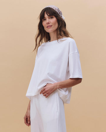 A woman with long brown hair wears THE GREAT. PF25 HEAVY JERSEY KNITS' The Track Crew in True White and pants, styled with a patterned headscarf for a vintage athletic look against a beige background.
