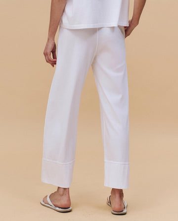 Person stands on a beige surface, seen from the back, wearing THE GREAT. PF25 HEAVY JERSEY KNITS The Jersey Win Pant in True White with a white top and white slide sandals for an easy loungewear look.
