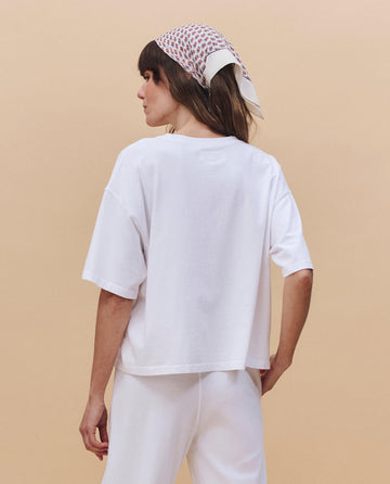 A person stands with their back to the camera, wearing THE GREAT. PF25 HEAVY JERSEY KNITS "The Track Crew." in True White, paired with white pants and a patterned headscarf, set against a beige background.