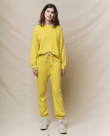 A person in a Daffodil-colored The Teammate Sweatshirt by THE GREAT. SP25 KNITS and matching sweatpants with white sneakers stands against a plain backdrop, effortlessly embodying casual apparel.