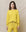 A person stands against a beige backdrop, donning The College Sweatshirt in Solid Daffodil by THE GREAT. SP25 KNITS, along with matching pants, presenting a fashionable twist on collegiate style.