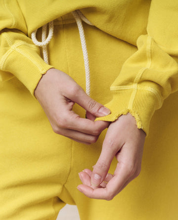 Close-up of a person in THE GREAT. SP25 KNITS The College Sweatshirt in Daffodil, adjusting the frayed sleeve edge with both hands. A white drawstring peeks from their collegiate clothing pants.