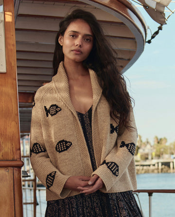 A woman with long hair stands on a boat, wearing THE GREAT. SU26 The Swimming Fish Lodge Cardigan in Natural over a dark patterned dress, with water and trees in the background.