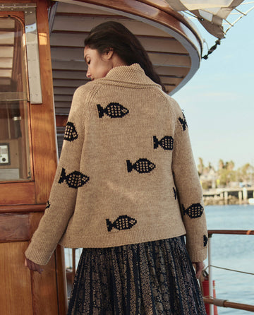 A woman stands on a boat deck wearing THE GREAT. SU26's The Swimming Fish Lodge Cardigan in Natural, featuring black fish patterns, paired with a patterned skirt. Water and palm trees fill the background.
