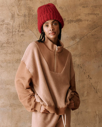 A person wearing The Handknit Chunky Beanie in Red Berry from THE GREAT. HOL 24 PLUSH KNITS and a tan zip-up fleece stands against a textured beige wall.