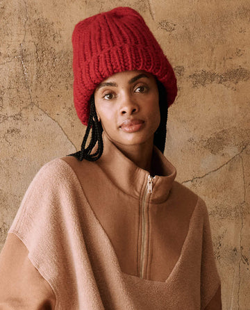 A person is sporting "The Handknit Chunky Beanie" in Red Berry by THE GREAT. HOL 24 PLUSH KNITS, paired with a brown zip-up sweater, and is standing against a textured, beige wall.