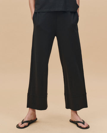 Wearing THE GREAT. PF25 HEAVY JERSEY KNITS The Jersey Win Pant in Almost Black with black flip-flops, standing against a plain beige background. Upper body is partially out of frame.