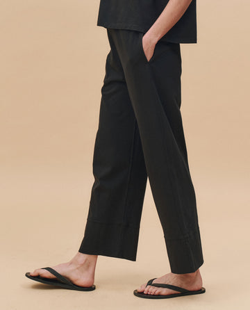 Wearing THE GREAT. PF25 HEAVY JERSEY KNITS The Jersey Win Pant in Almost Black with a matching top and black flip-flops, this person stands with one hand in their pocket against a neutral beige background.