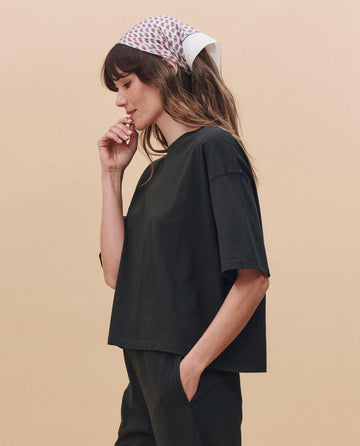 A woman stands in profile against a beige background, wearing THE GREAT. PF25 HEAVY JERSEY KNITS The Track Crew. -- Almost Black, matching pants, and a patterned headscarf, with one hand in her pocket and the other near her face.