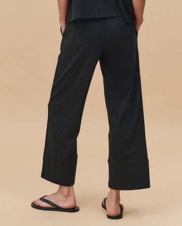 A person stands on a neutral background, shown from the back, wearing THE GREAT. PF25 HEAVY JERSEY KNITS The Jersey Win Pant in Almost Black and black flip-flops.