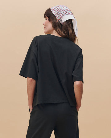 A person wearing THE GREAT.'s The Track Crew. -- Almost Black from the PF25 HEAVY JERSEY KNITS collection stands with their back to the camera, paired with matching pants and a patterned scarf tied around their head.