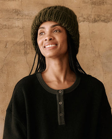 A person wearing "The Handknit Chunky Beanie" in Winter Moss from THE GREAT. HOL 24 PLUSH KNITS and a dark henley shirt smiles while looking off to the side, with a textured wall as the background.
