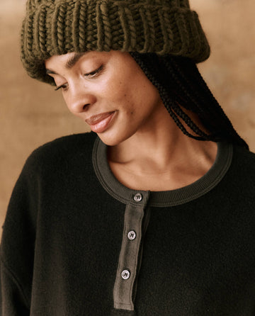 The individual, sporting The Handknit Chunky Beanie in Winter Moss from THE GREAT. HOL 24 PLUSH KNITS and braids, gazes downward with a subtle smile.