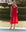 A person in THE GREAT. SP25 SWIM The Ruffle Coverup—Hot Red stands barefoot by the pool with manicured hedges behind, effortlessly showcasing stylish women's beachwear.