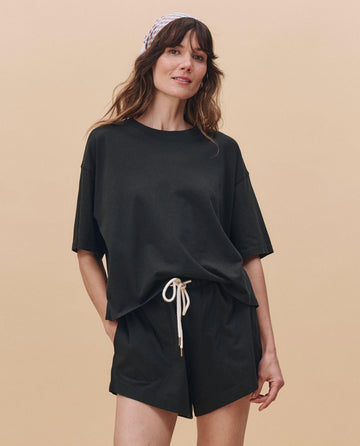 A woman stands against a beige background in THE GREAT. PF25 HEAVY JERSEY KNITS The Flutter Short in Almost Black, styled with an oversized t-shirt and patterned headscarf for an effortlessly chic, relaxed look made in Los Angeles.