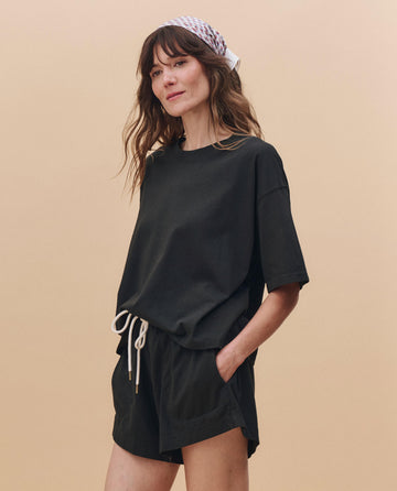 A woman stands against a beige background wearing THE GREAT. PF25 HEAVY JERSEY KNITS The Flutter Short in Almost Black, a black t-shirt, and a white patterned headscarf.