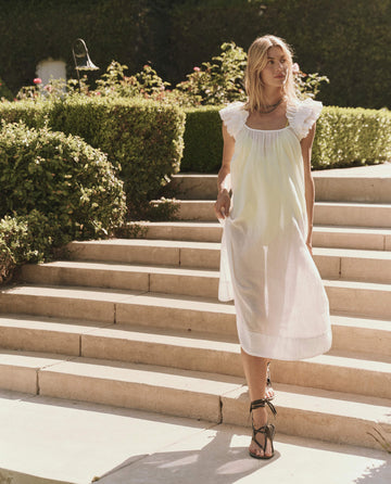 A woman wearing THE GREAT. SP25 SWIM's The Ruffle Coverup in white with ruffle shoulders walks down sunlit stone steps, embracing effortless fashion.