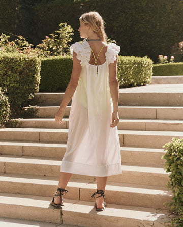 A woman in THE GREAT. SP25 SWIM's The Ruffle Coverup—White, a sleeveless midi dress, stands on outdoor stone steps amid lush greenery for an effortlessly stylish look.