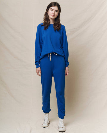 Someone in The College Sweatshirt, Solid -- Bright Cobalt by THE GREAT. SP25 KNITS and matching sweatpants stands against a beige fabric background, wearing white sneakers.