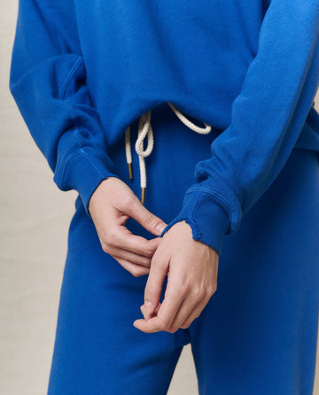 A person wearing The College Sweatshirt in Bright Cobalt by THE GREAT. SP25 KNITS, featuring white drawstrings, reminiscent of college apparel, with hands clasped in front.