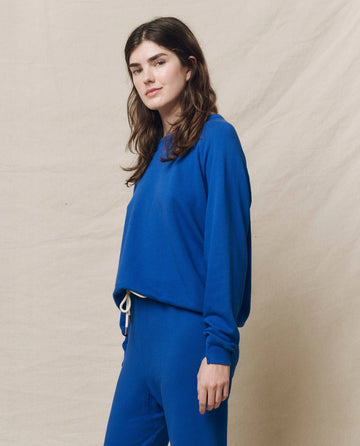 A person dressed in The College Sweatshirt by THE GREAT. SP25 KNITS in Bright Cobalt stands against a beige backdrop, facing slightly sideways.