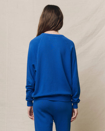 A person with long brown hair, wearing THE GREAT. SP25 KNITS' "The College Sweatshirt" in Bright Cobalt and pants, stands with their back to the camera against a neutral background.