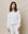 A woman with long hair stands against a plain backdrop, wearing The College Sweatshirt in True White and matching pants by THE GREAT. SP25 KNITS, crafted from double-washed terry.