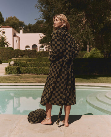 A person in THE GREAT. SP24 SWIM The Terry Robe – Dark Army Check stands barefoot by a pool, holding a matching bag, with a house and greenery in the background.