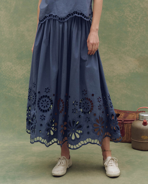 Only the lower body is visible as a person stands on a greenish surface wearing THE GREAT. SU25’s The Super Bloom Eyelet Skirt in Vintage Blue, featuring laser-cut floral details at the hem, paired with white sneakers and a vintage picnic set nearby.