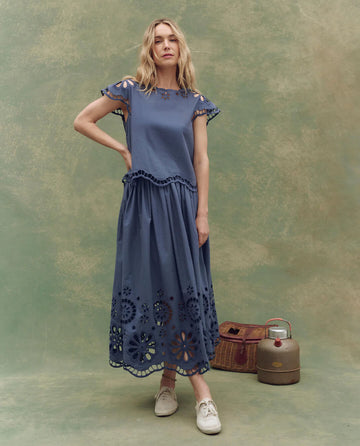 A woman stands against a green backdrop wearing THE GREAT. SU25 The Super Bloom Eyelet Skirt in Vintage Blue, paired with white sneakers, next to a vintage basket and metal canister.