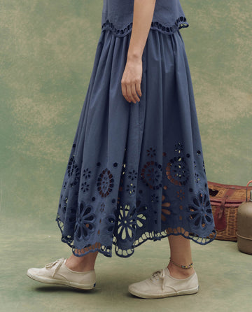 A person in THE GREAT. SU25's The Super Bloom Eyelet Skirt—Vintage Blue—and beige sneakers stands on a green background with a picnic basket nearby, capturing the essence of a Super Bloom afternoon.