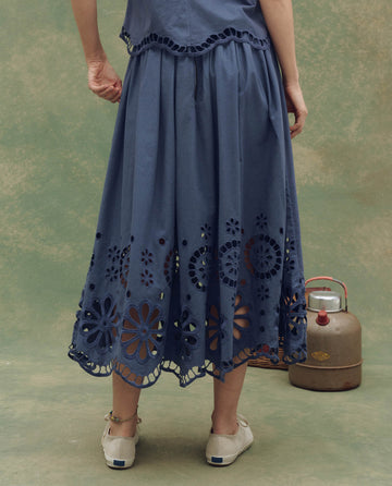 A person stands on a greenish floor wearing THE GREAT. SU25 The Super Bloom Eyelet Skirt in Vintage Blue with beige shoes, while an old-fashioned metal kettle sits in the background.