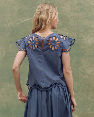A woman faces away, wearing THE GREAT. SU25's The Super Bloom Eyelet Top in Vintage Blue, featuring cut-out flower patterns on the sleeves and back, paired with a matching skirt against a muted green background.