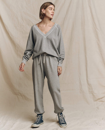 A person stands in front of a beige backdrop, wearing The V-Neck Sweatshirt in Varsity Grey by THE GREAT. FALL 24 KNITS, paired with matching pants and sneakers, exuding a touch of casual elegance with a nod to vintage wash style. Their relaxed outfit epitomizes the laid-back vibe reminiscent of Los Angeles fashion.