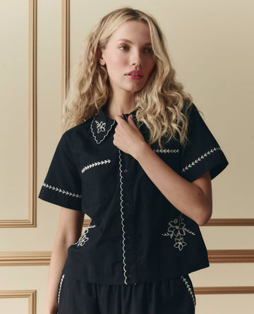 A woman with long blonde hair models THE GREAT. SP26 D2 The Bowling Shirt W/ Hanky Embroidery in black, featuring short sleeves and white hanky embroidery, against a beige paneled background.
