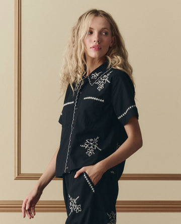 A woman with wavy blonde hair wears THE GREAT. SP26 D2's The Bowling Shirt W/ Hanky Embroidery in black, styled with matching pants, standing against a beige wall with decorative molding.