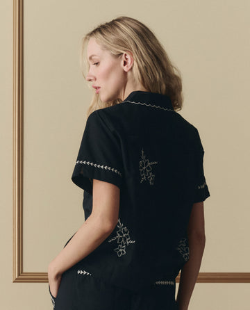 A woman with long blonde hair stands in profile against a beige background, wearing THE GREAT. SP26 D2's The Bowling Shirt W/ Hanky Embroidery in black, featuring white embroidered details and short sleeves.