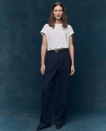 A woman faces forward against a blue textured backdrop, wearing THE GREAT. CORE KNITS The Boxy Crew. Solid -- True White t-shirt, paired with a black belt, dark pinstripe trousers, and black shoes.