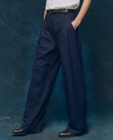 A person wearing THE GREAT. F25 COLLECTORS The Menswear Trouser in Dark Blue Stripe, paired with a white shirt, black leather belt, and black shoes, stands against a dark textured background.