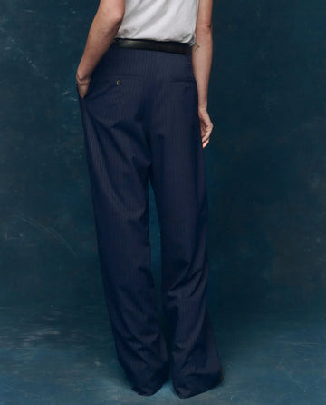 Person standing with their back to the camera, wearing THE GREAT. F25 COLLECTORS The Menswear Trouser – Dark Blue Stripe, paired with a light grey shirt and set against a dark blue backdrop.